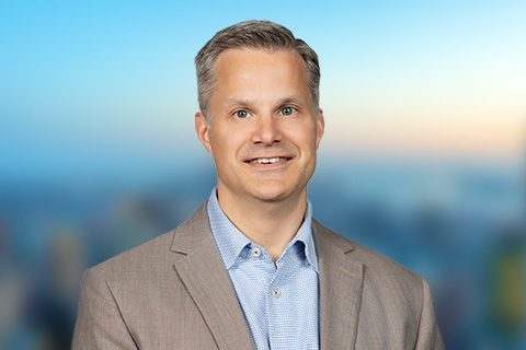 Man in a beige blazer smiles in front of a bright window with a blurred city view.