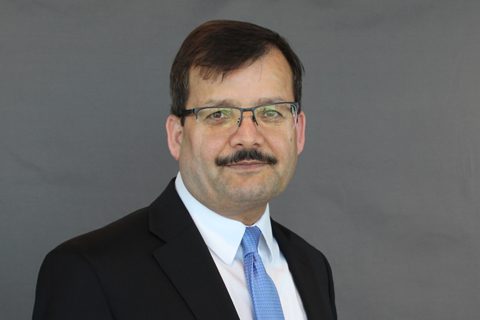 A man wearing glasses, a dark suit, a white shirt, and a light blue tie stands in front of a plain gray background.