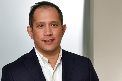 A man wearing a dark suit jacket and white collared shirt stands indoors in front of a light-colored background, facing the camera and smiling slightly.