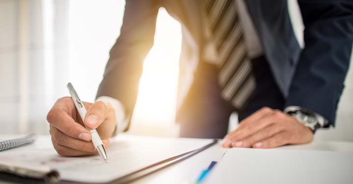 A person in a suit writes on a document at a desk, holding a pen in one hand while leaning on the table with the other.