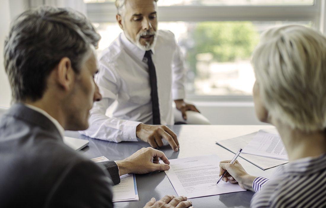Three professionally dressed people sit at a table discussing and reviewing documents, with one person signing a paper while another points at it.