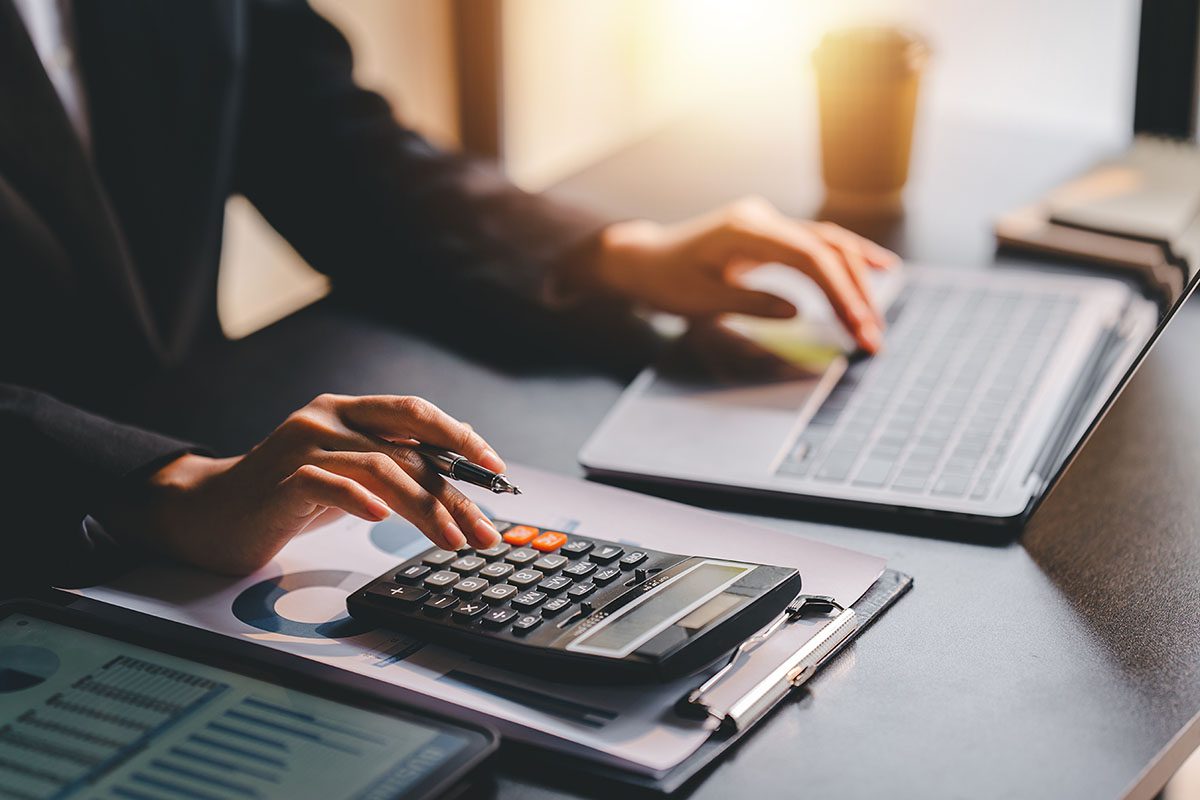 Person working at a desk using a laptop and calculator, with financial documents and graphs visible on the desk.