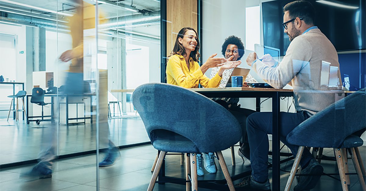 Three people sit at a table in a modern office meeting room, engaged in conversation, while a person walks past outside the glass walls.