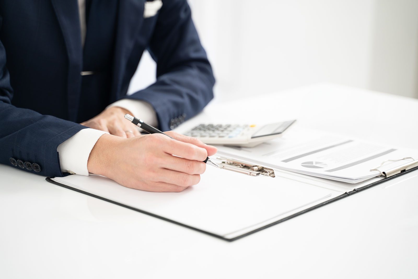 A person in a suit writes on a clipboard at a desk with documents, a calculator, and a pen in view.