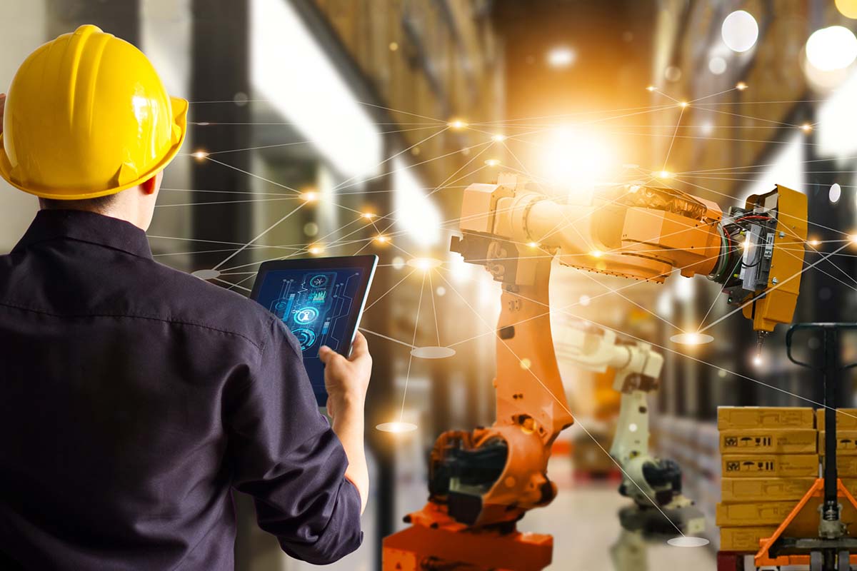 A person wearing a yellow hard hat uses a tablet to control an industrial robotic arm in a factory setting with digital network graphics overlay.