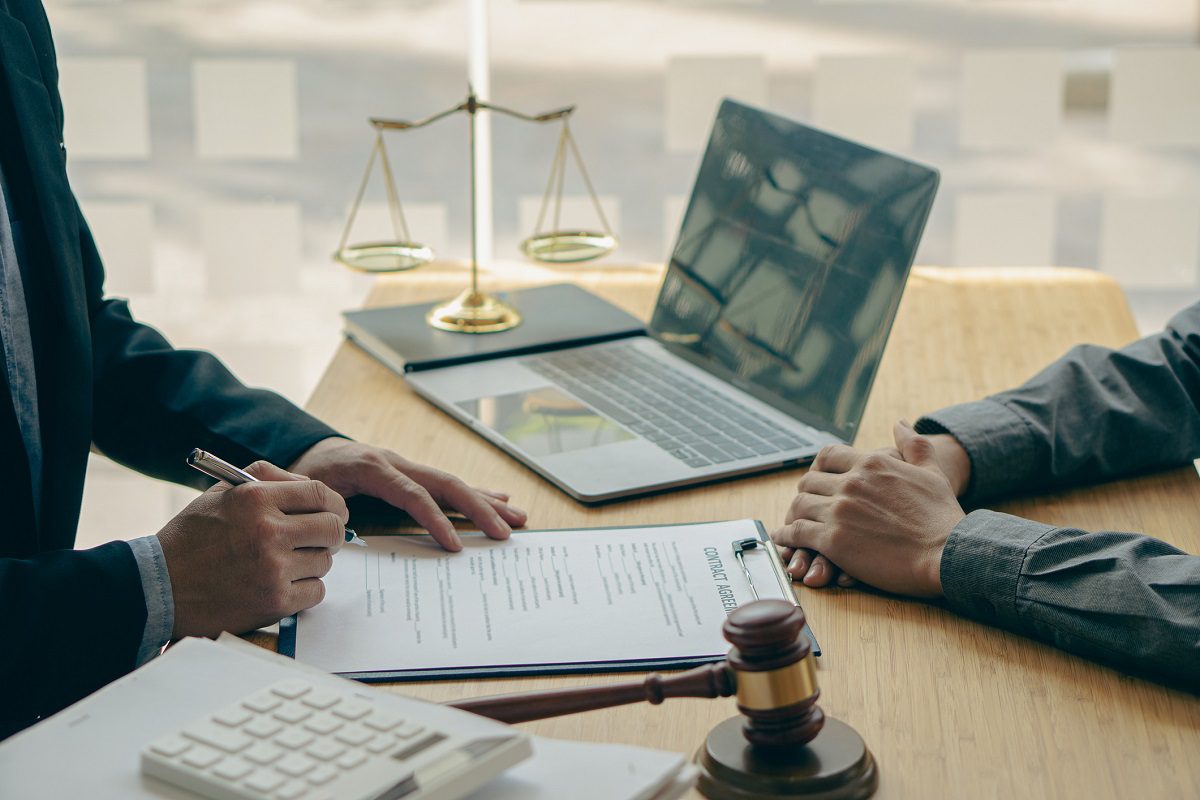 Two people sit at a desk with legal documents, a laptop, a gavel, a scale of justice, and a calculator; one person is signing a document.