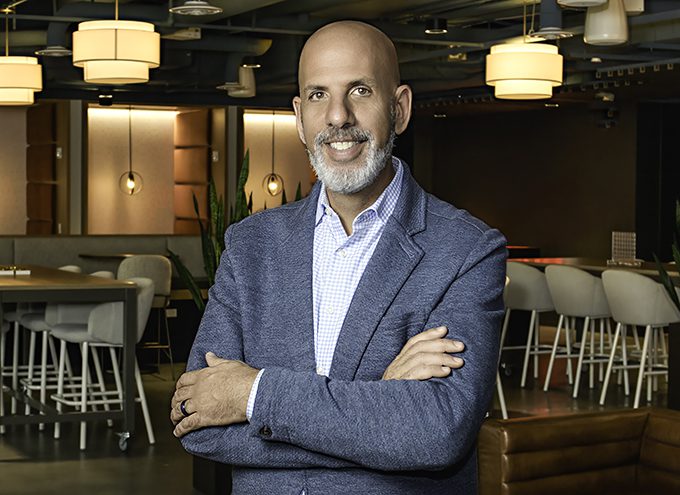 A man with a bald head and gray beard, wearing a blue blazer and checkered shirt, stands with arms crossed in a modern, well-lit office space.