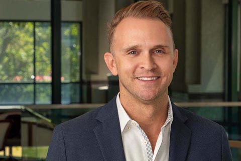 A man in a dark suit jacket and white shirt stands indoors, smiling, with large windows and greenery visible in the background.