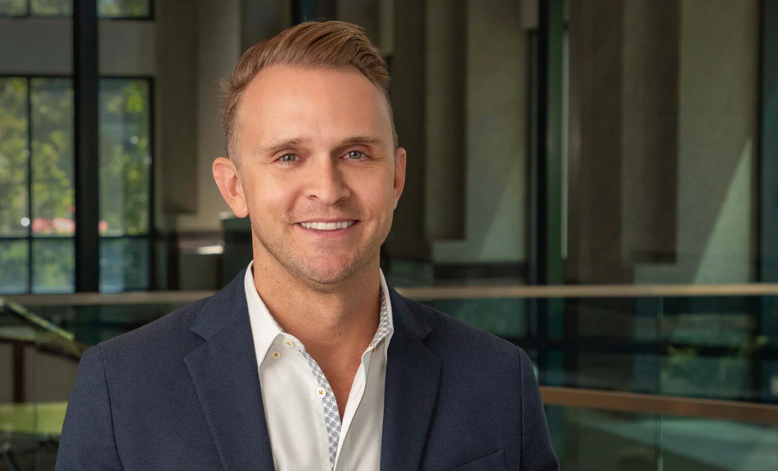 A man in a navy blazer and white shirt smiles while standing indoors in front of large windows and glass railings.