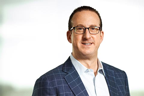 A man wearing glasses and a blue plaid suit jacket over a white shirt, smiling, with a blurred light background.