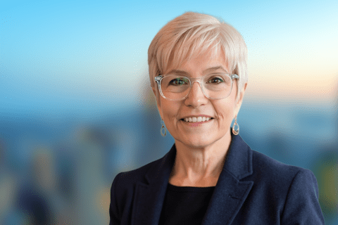 A woman with short gray hair and glasses, wearing a navy blazer, smiles at the camera. The background is blurred with soft blue and orange tones.