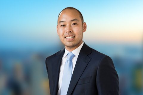 Man in a black blazer smiles in front of a bright window with a blurred city view.