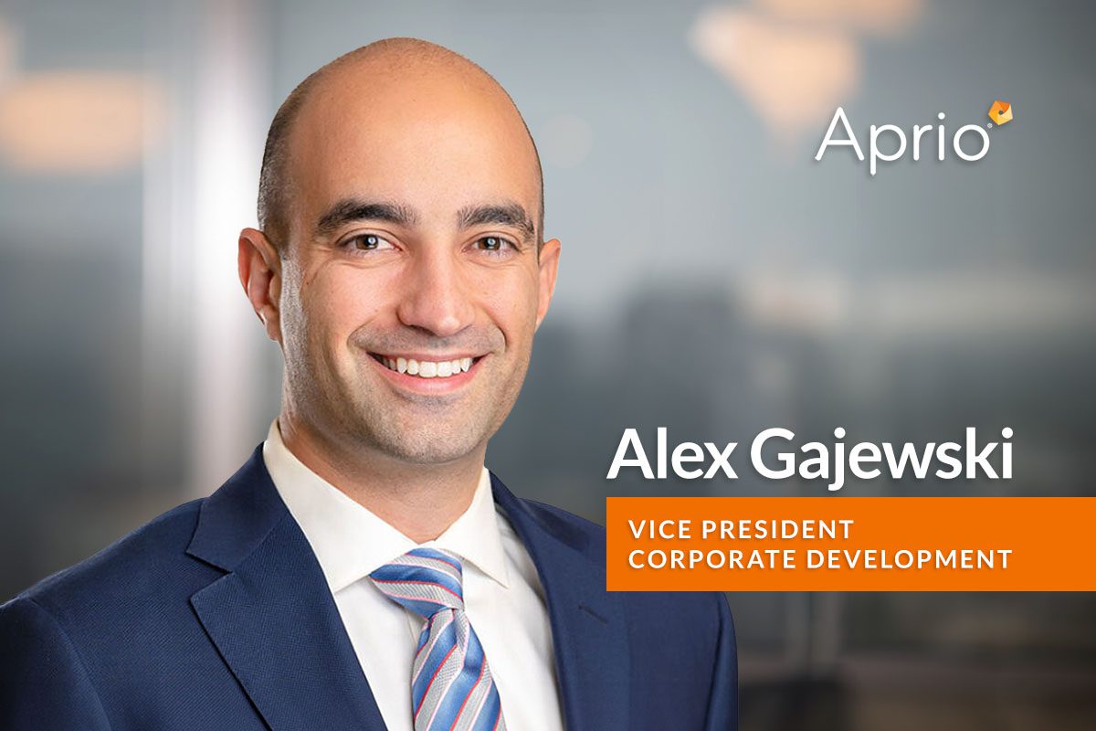A man in a suit and tie smiles at the camera; text reads Alex Gajewski, Vice President, Corporate Development, Aprio, with a blurred office background.