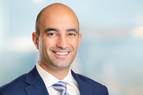 A man in a blue suit and striped tie smiles at the camera against a blurred light blue background.