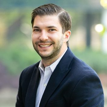 A man with short brown hair and a beard, wearing a dark blazer and light shirt, smiles at the camera outdoors with greenery in the background.