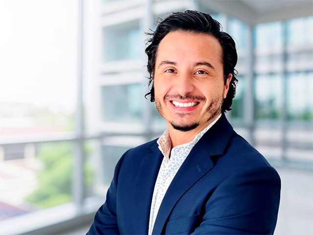 A man with dark hair and a beard, wearing a navy suit and patterned shirt, smiles in a modern office setting with large windows.