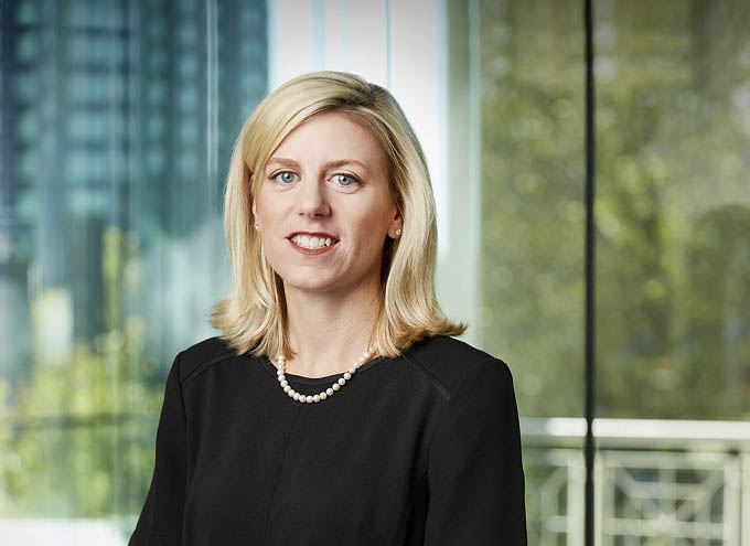 A woman with shoulder-length blonde hair wearing a black top and pearl necklace stands indoors with large windows and greenery in the background.
