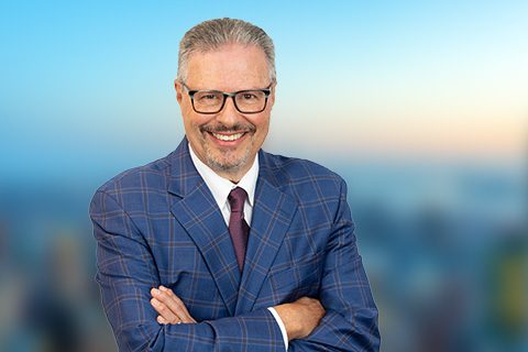 A man with gray hair and glasses, wearing a blue plaid suit, white shirt, and maroon tie, stands with arms crossed, smiling against a blurred cityscape background.