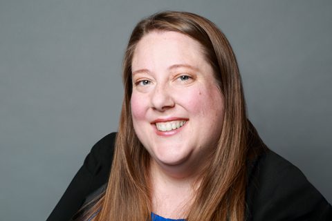 A woman with long straight brown hair, wearing a blue top and black jacket, smiles at the camera against a plain gray background.