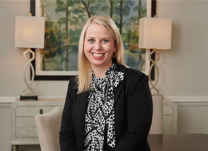 A woman with straight blonde hair, wearing a black blazer and patterned blouse, sits and smiles in a well-lit office with lamps and artwork in the background.