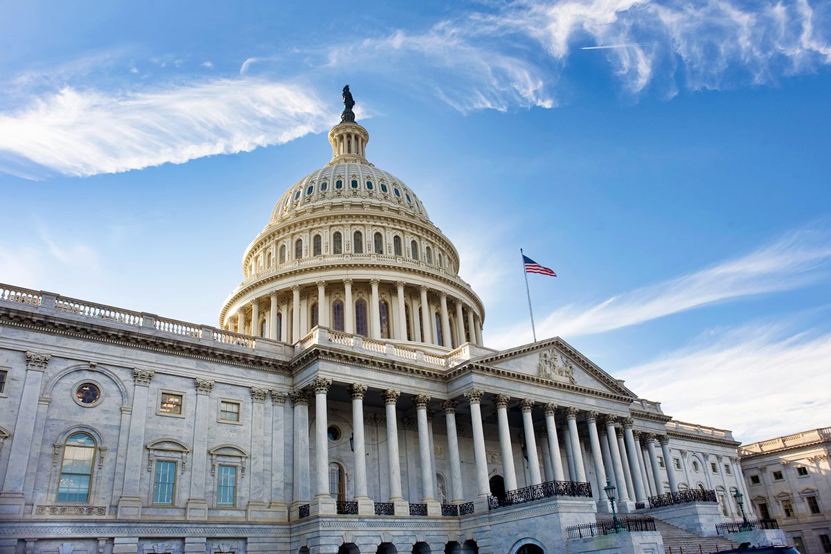 The United States Capitol building with its dome, columns, and an American flag, set against a blue sky with wispy clouds.