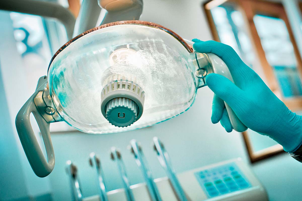 A gloved hand adjusts a dental overhead light in a dentist’s office, with dental instruments visible in the background.