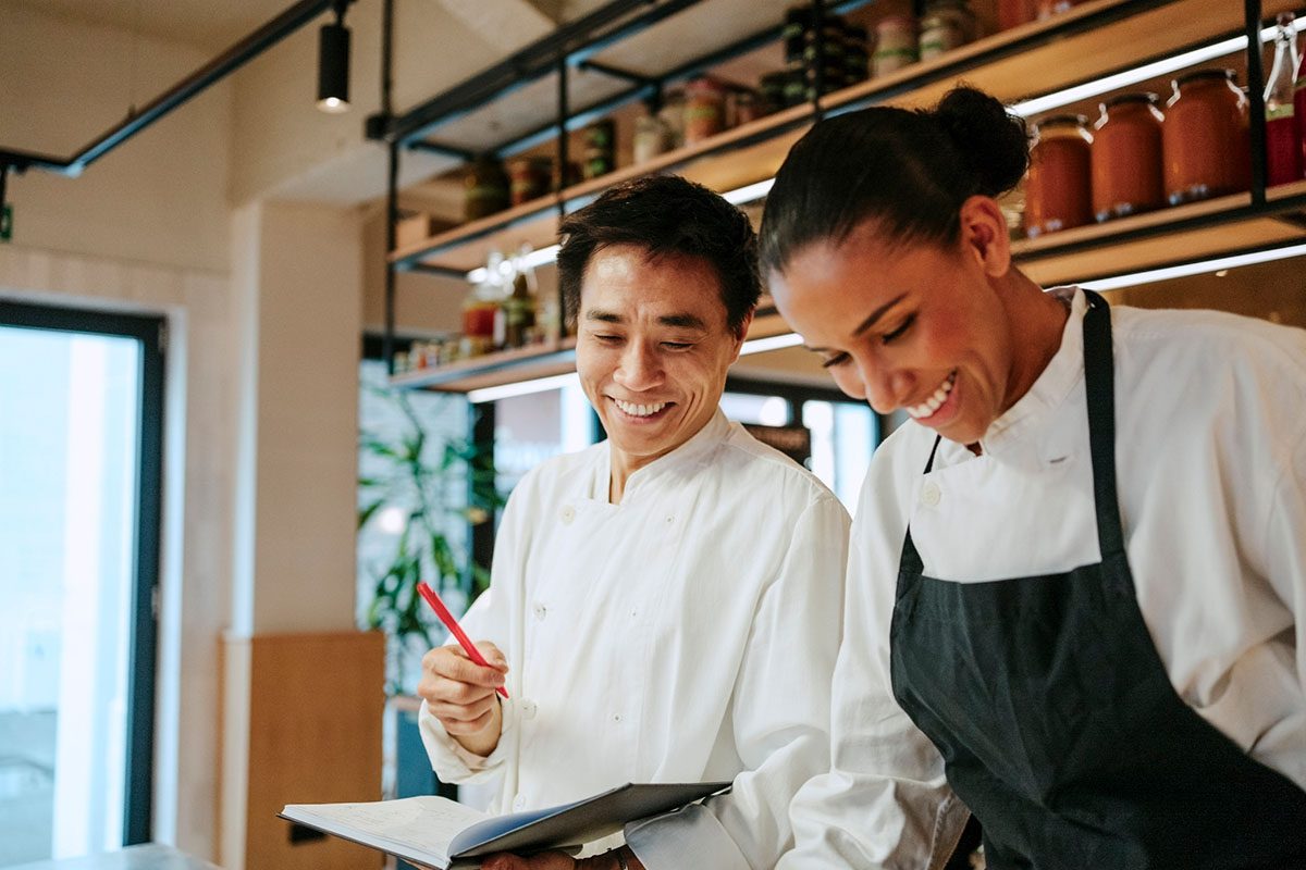 Two chefs in a kitchen, one holding a pen and notebook, both smiling and wearing white uniforms. Shelves with jars are visible in the background.