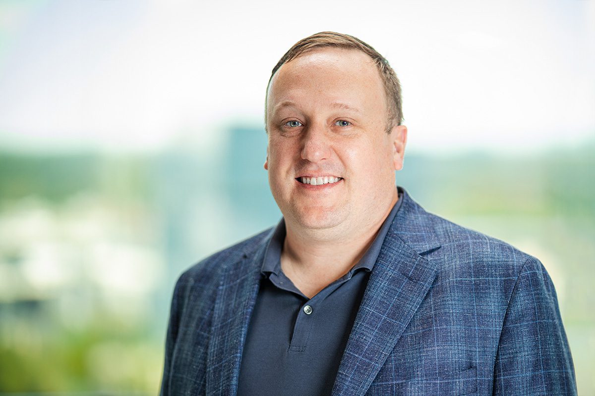 A man in a blue blazer and dark polo shirt stands indoors, smiling, with a blurred outdoor background behind him.
