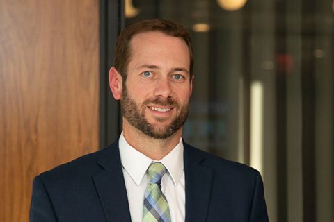 A man with a beard in a suit and striped tie stands indoors near a wooden wall and glass panels.