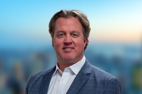 Man in a blue blazer smiles in front of a bright window with a blurred city view.