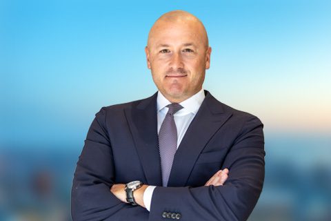 A bald man in a dark suit, white shirt, and striped tie stands with arms crossed against a blurred outdoor backdrop.