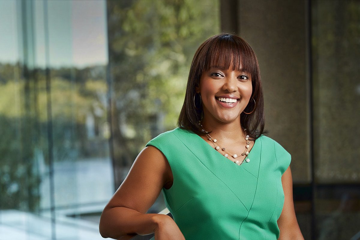 A woman in a green dress smiles at the camera while standing indoors near a large window with greenery outside.