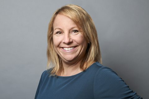 A woman with blonde hair, wearing a blue top, smiles at the camera against a plain gray background.