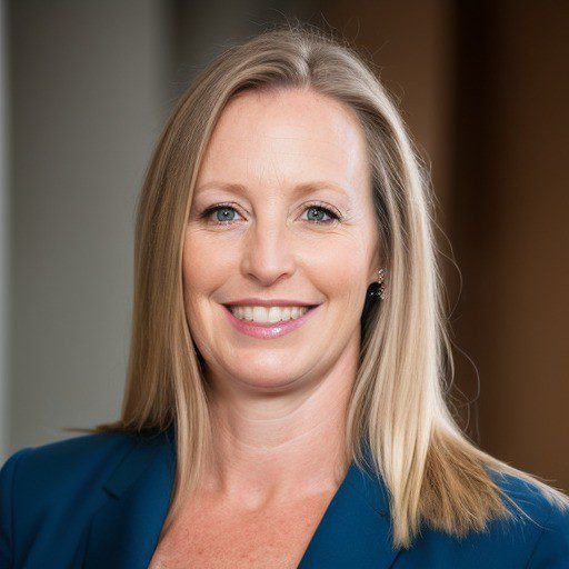 A woman with straight blonde hair, wearing a blue blazer, smiles at the camera against a blurred indoor background.