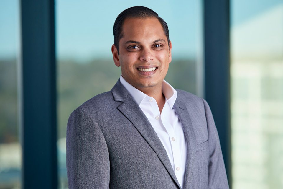 A man in a gray suit and white shirt stands indoors, smiling at the camera with large windows and a blurred outdoor background behind him.