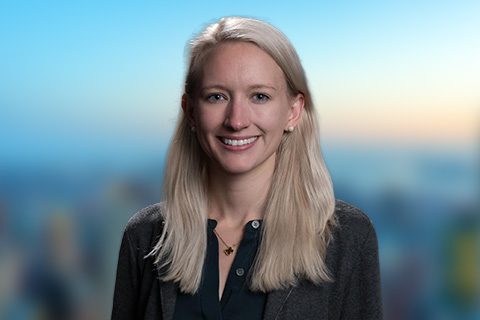 Blonde woman in a black sweater smiles in front of a bright window with a blurred city view