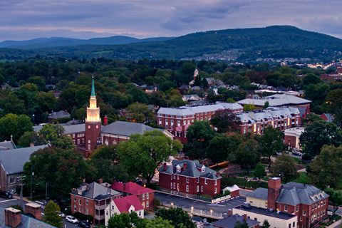 Aerial view of a small town with a church steeple, brick buildings, and tree-lined streets, set against a background of hills under a cloudy sky.