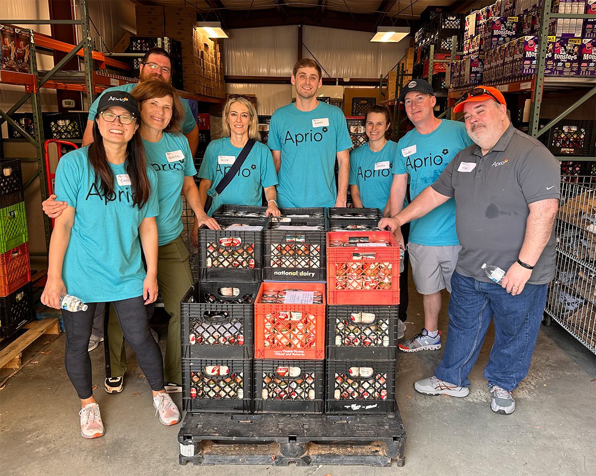 A group of eight people wearing Aprio shirts stand in a warehouse behind stacked crates of packaged food.
