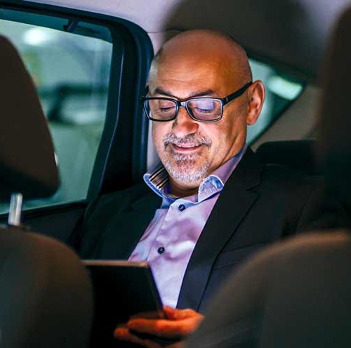 Bald man with glasses sitting in the back seat of a car, looking at a tablet and smiling.