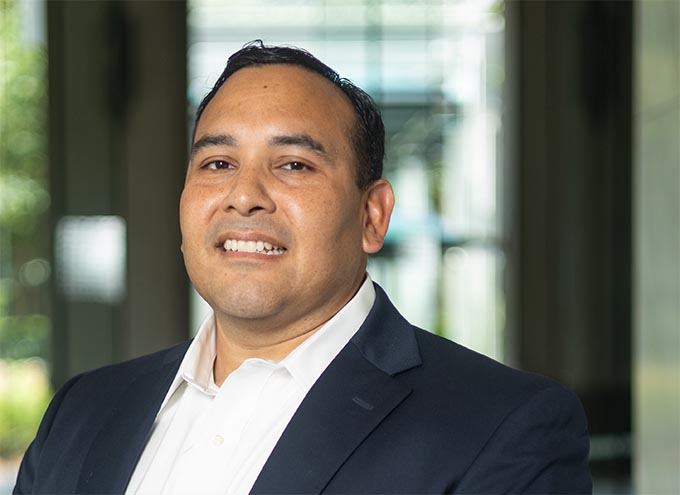 A man wearing a dark suit jacket and white shirt smiles slightly while standing indoors with a blurred background.