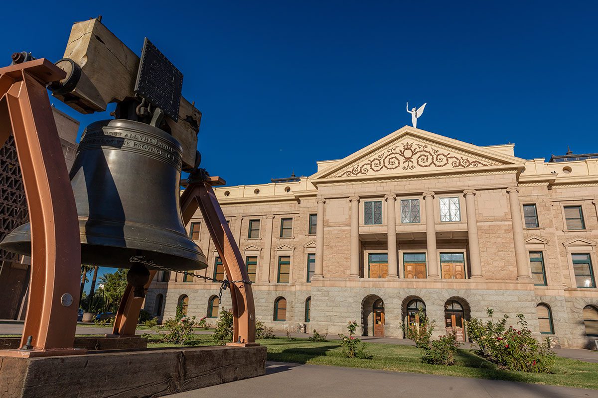 A large bell is displayed in front of a neoclassical government building with columns and a statue on the roof under a clear blue sky.