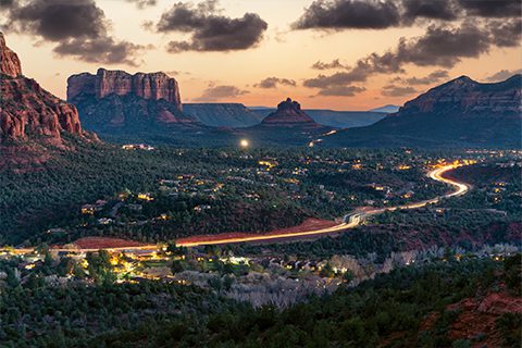 A winding road with car lights runs through a valley surrounded by red rock formations and dense greenery at sunset, under a partly cloudy sky.