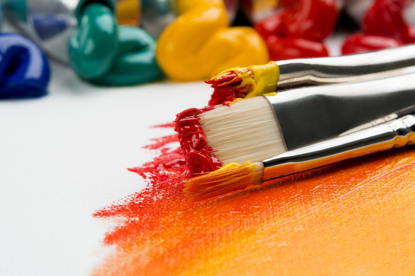 Two paintbrushes coated in red and yellow paint rest on a canvas with red and orange paint strokes, with colorful paint tubes in the background.
