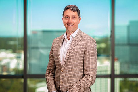 A man in a checked beige blazer and white shirt stands smiling in front of a large window with a cityscape in the background.