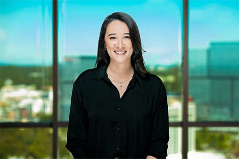 A woman with long dark hair wearing a black shirt stands indoors in front of large windows overlooking a cityscape.