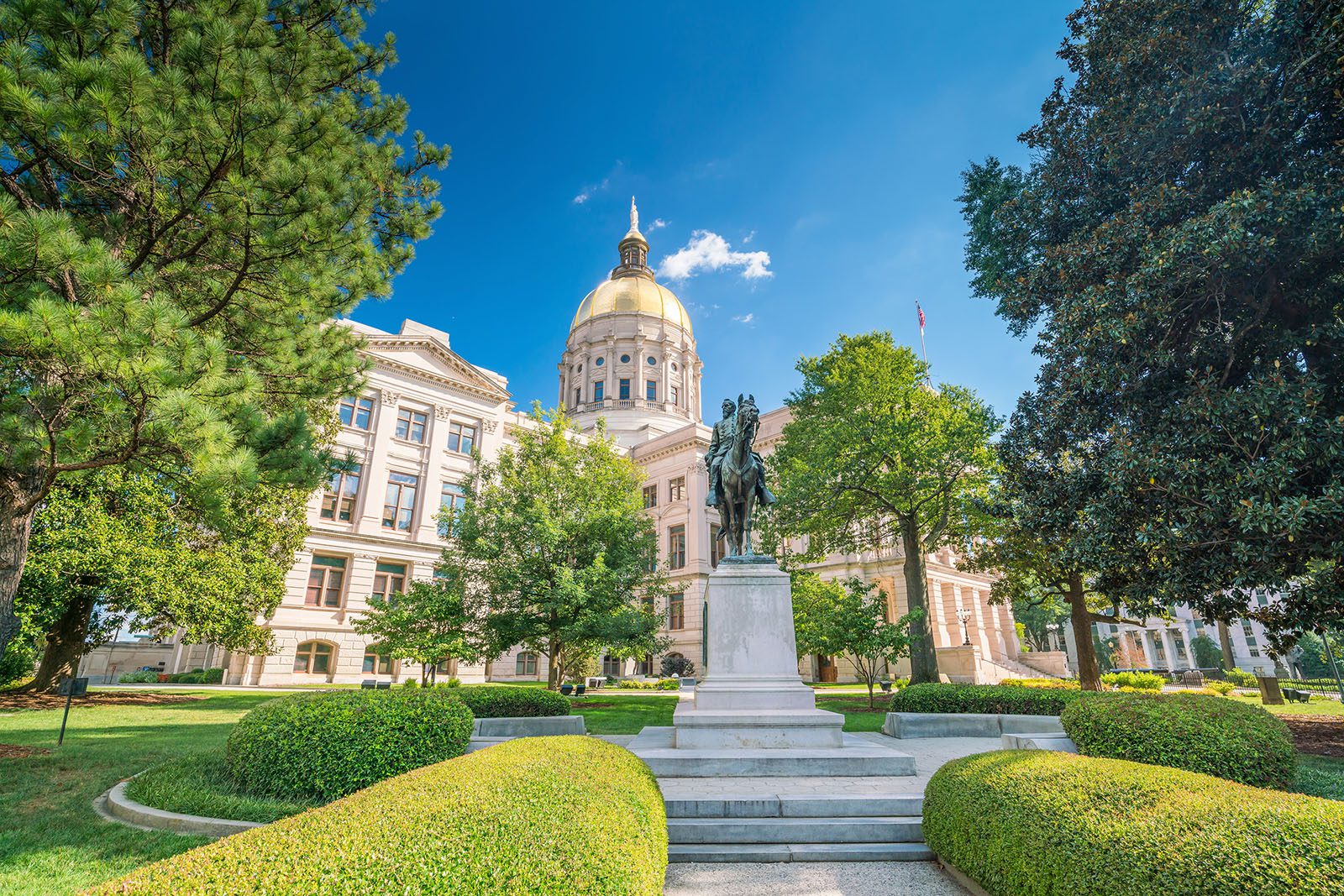 A domed government building with a gold roof stands behind a statue surrounded by manicured trees and bushes under a clear blue sky.