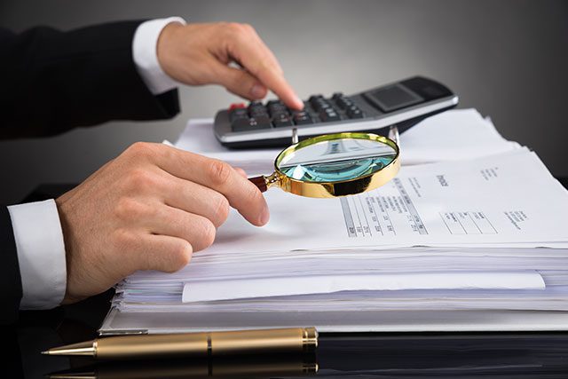 A person examines financial documents with a magnifying glass while using a calculator; a pen rests on the desk nearby.