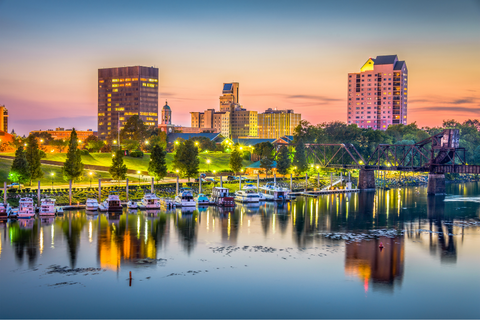 City skyline at sunset with tall buildings, waterfront trees, boats docked along the river, and their reflections in the water.