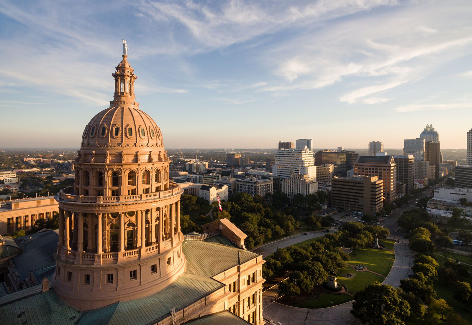 Aerial view of the Texas State Capitol building with downtown Austin city skyline in the background under a partly cloudy sky.
