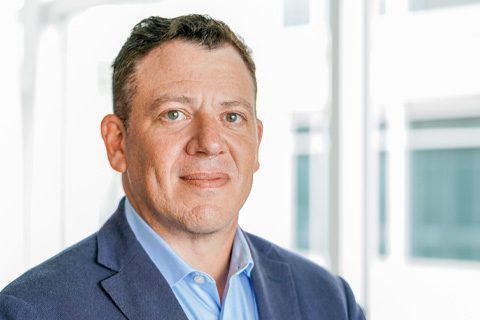 A man in a blue blazer and light blue shirt stands indoors in front of large windows with a neutral expression.